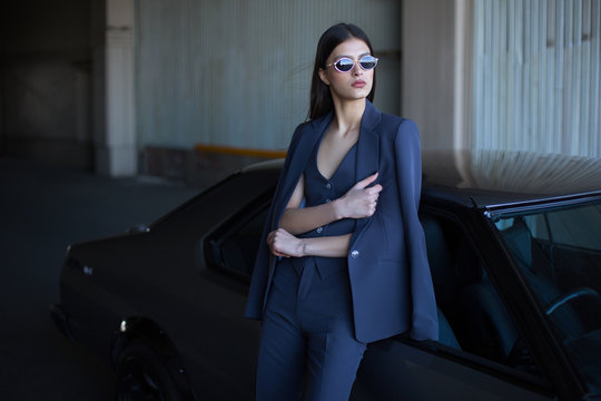 Mafia lady outside japonese car in the sea port. Fashion girl standing next to a retro sport car on the sun. Stylish woman in a blue suit and sunglasses waiting near classic car.