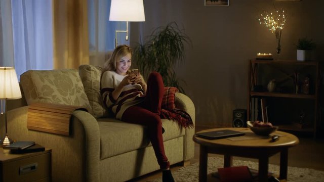Beautiful Young Woman Enters Living Room in the Evening. Sits on Her Sofa and Starts Using Smartphone. Shot on RED EPIC-W 8K Helium Cinema Camera.