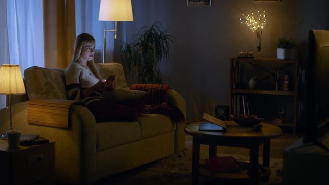 In the Evening Beautiful Young Woman Relaxes on a Couch in Her Cozy Living Room. She Uses Her Smartphone. Shot on RED EPIC-W 8K Helium Cinema Camera.