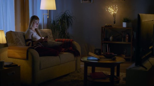In the Evening Beautiful Young Woman Relaxes on a Couch in Her Cozy Living Room. She Uses Her Smartphone and Simultaneously Watches TV. Shot on RED EPIC-W 8K Helium Cinema Camera.