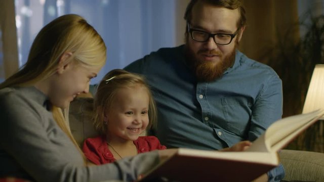 Father, Mother and Little Daughter Reading Children's Book on a Sofa in the Living Room. It's Evening. Shot on RED EPIC-W 8K Helium Cinema Camera.