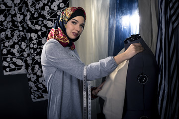 Female fashion designer tailoring conservative clothing in a textile workshop.  The hijab she is...