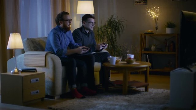 In the Evening Two Friends are Sitting on a Sofa in the Living Room and  Playing Comptetitive Video Games. Shot on RED EPIC-W 8K Helium Cinema Camera.