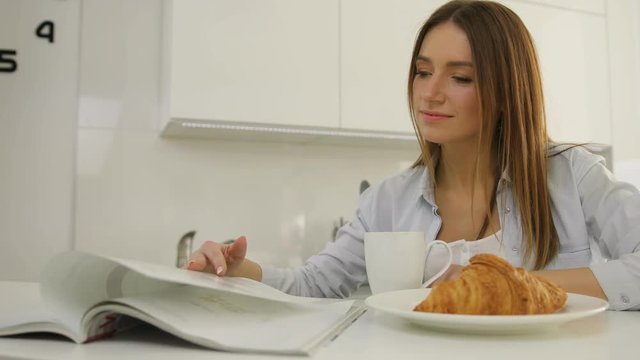 Young woman thumbing through the magazine in the kitchen during breakfast