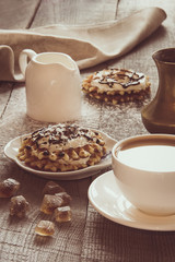 White cup of coffee with milk, brown sugar and homemade waffle with whipped cream with decor on top. Tasty breakfast. Vintage toned.