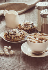 White cup of coffee with milk, marshmallow and homemade waffle with whipped cream with decor on top. Tasty breakfast. Vintage toned.