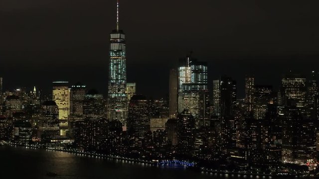 AERIAL HELI SHOT: Skyline view of New York peninsula iconic Downtown Manhattan financial district with famous huge waterfront skyscrapers and giant office buildings lit up with lights shining at night