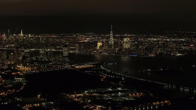 AERIAL HELI SHOT: Flying above New Jersey port in industrial zone at night, overlooking iconic NYC Downtown skyline with brightly lit skyscrapers WITH Brooklyn and Queens cityscape in the background