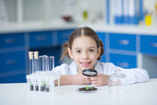 Little girl scientist holding magnifier and working with green plant in soil