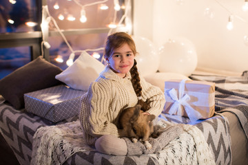 Very beautiful and charming little dark-haired girl in white sweater holds a live rabbit in the interior of the house. New year. Christmas. Hare. Studio.
