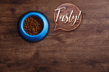 Bowl of dry kibble dog food. Tasty speech bubble. Healthy pets feed. Blue plate on wooden rustic background.