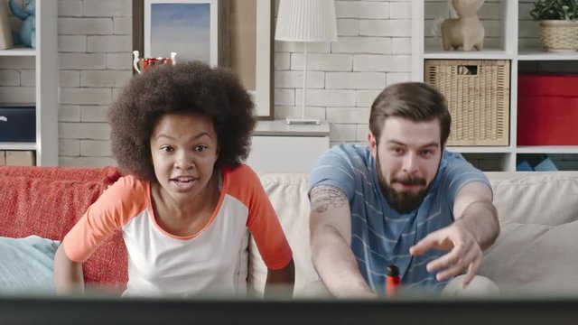 Young bearded man and teenage African girl watching live match on TV with excitement