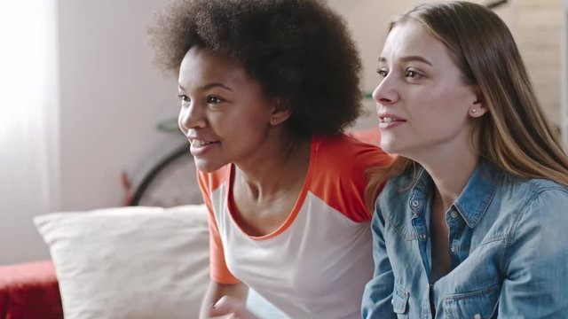 Closeup of African and Caucasian teen girls watching sports match on TV at home. They shouting at screen and embracing while celebrating victory of favorite team