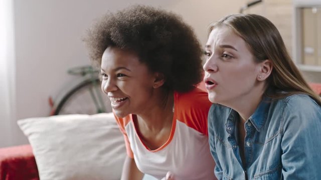 Closeup of African and Caucasian young women watching sports match on TV at home. They yelling at screen and raising hands while celebrating goal