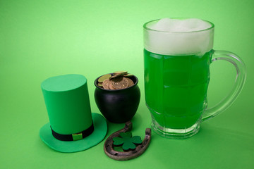 St Patrick's Day green beer with shamrock, pot with gold coins, horseshoe and Leprechaun hat against green background.