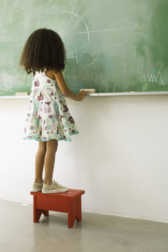 Little girl standing on stool in front of blackboard, rear view