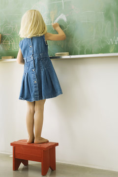 Little girl standing on stool, scribbling on blackboard, rear view