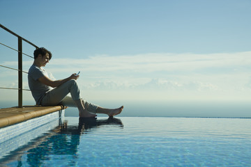 Man sitting on edge of infinity pool, text messaging with cell phone