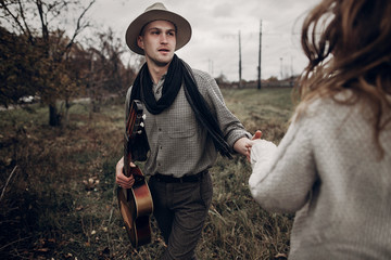 Romantic hipster couple, handsome musician man with guitar and gypsy woman in boho sweater dancing in autumn field - obrazy, fototapety, plakaty