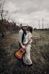 Stylish hipster couple hug in field, handsome cowboy musician with guitar and indie style woman in white sweater pose near railway station