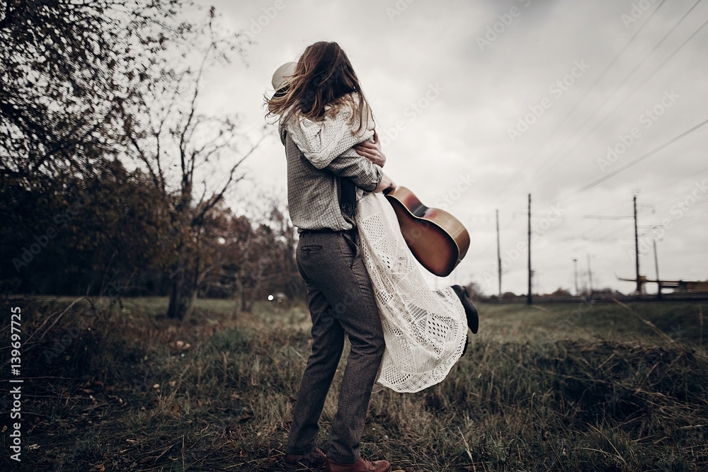 Wall mural stylish hipster couple dancing in windy field. boho gypsy woman and man in hat embracing in windy fi