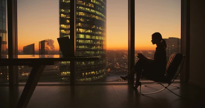young woman businessman ceo sitting in a chair with a cup of coffee in the hands of the panoramic window in the office skyscrapers at sunset