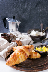 Breakfast with two croissant, butter, cup of coffee, cottage cheese and sliced mango fruit, served on serving wood board with textile napkin on white and gray concrete texture background. Close up.