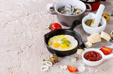 Breakfast with fried quail eggs in iron cast pan, cherry tomatoes, onion, ketchup sauce, seasonings in mortar, cheese grater, egg shell in pot. Beige concrete texture background. Copy space.