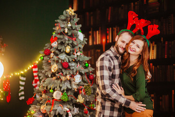 Young family with deer antlers on their heads hugs before Christmas tree