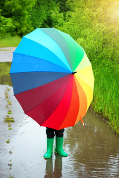 Child walking in wellies in puddle on rainy weather. Boy holding colourful umbrella under rain in summer