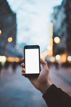 Vertical image of male hand holding smartphone with blank screen with copy space for your design or logo, mockup of black cellphone, evening city street on the background, bokeh light