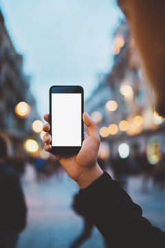 Vertical image of young hipster man holding smartphone with blank screen with area for your design or logo, mock up of modern black cellphone, evening city street on the background with bokeh lights