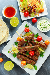 Homemade Kofta kebabs on skewers with pita, lime, vegetables, sweet chili and yogurt sauce on white plate.