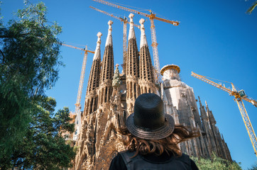 Young woman traveler in a hat looks at the Sagrada Familia the most famous tourist attraction of...
