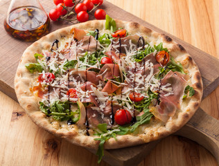 Pizza with dried ham, rocket leaves, tomato and Parmesan cheese on a cutting board.