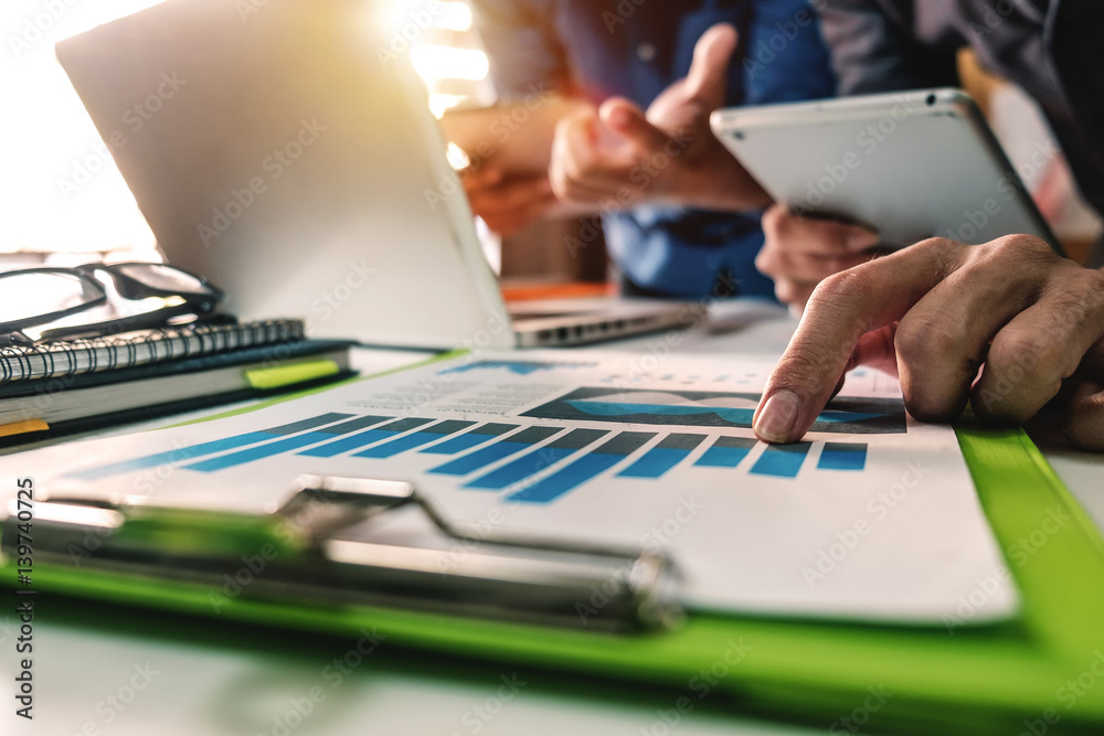 Wall mural businessman documents on office table with smart phone and laptop and two colleagues discussing data in the background in morning light