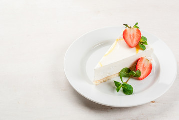 Light dessert: classic cheesecake, decorated with mint and strawberries. On a white plate, on a white wooden table. 