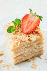 Light dessert: a classic Napoleon or millefeuille cake decorated with mint and strawberries. On a white plate, on a white wooden table. 