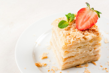 Light dessert: a classic Napoleon or millefeuille cake decorated with mint and strawberries. On a white plate, on a white wooden table. 