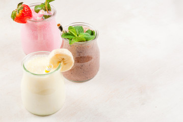 Selection of classic milkshakes in small jars: chocolate, strawberry, banana. On a white wooden table, with tubes, copy space