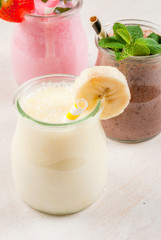 Selection of classic milkshakes in small jars: chocolate, strawberry, banana. On a white wooden table, with tubes, copy space