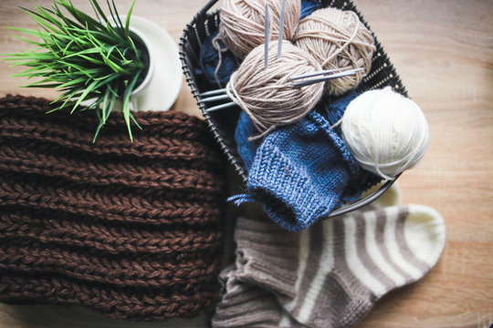Beige, white and blue yarn, knitting needles in the basket and a brown scarf. Striped beige-white knitted socks and a green plant in the pot. Wooden background. Knitting hobby