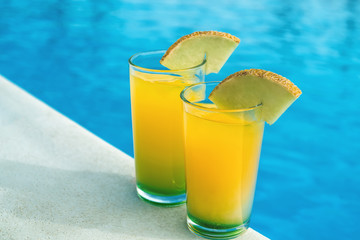 Glasses with yellow and green cocktail in front of a swimming pool with clear water. Close-up photo. Diagonal comprosition