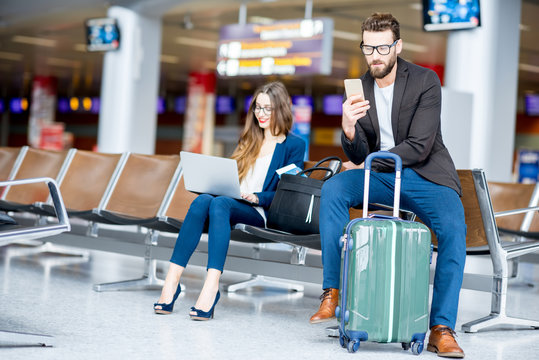 Elegant business couple sitting with laptop, phone and suitcase at the waiting hall in the airport. Business travel concept