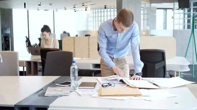 young working entrepreneur look for information among piles of papers, documents and folder for business analytics tasks staying at the table in the office open space during sunny day