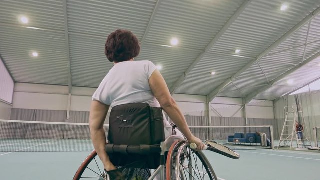 Disabled mature woman on wheelchair playing tennis on tennis court.