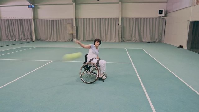 Disabled mature woman on wheelchair playing tennis on tennis court.