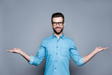 Smiling young man in jeans shirt showing a balance of two products isolated on gray background