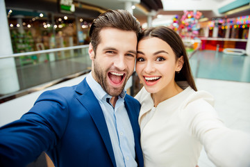 Fototapeta na wymiar Happy lovely young woman with her handsome boyfriend in suit together make selfie photo, walk in modern mall and having fun.