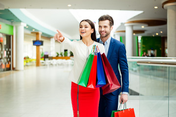 It's shopping time with  sales and fun. Cute cheerful  successful happy lovely couple in jacket holding colored shopping bags, pointing on shop  and laughing in mall at holiday